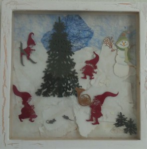 Nisser leger omkring et stort juletræ ude i naturen. Jule collage lavet af papir og i en træramme